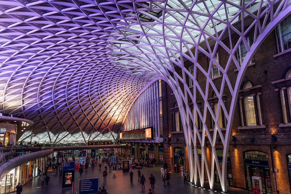 Inside a modern train station with a striking, architecturally intricate ceiling made of a lattice of white, curved steel beams illuminated by purple and white lighting. The station's interior feature